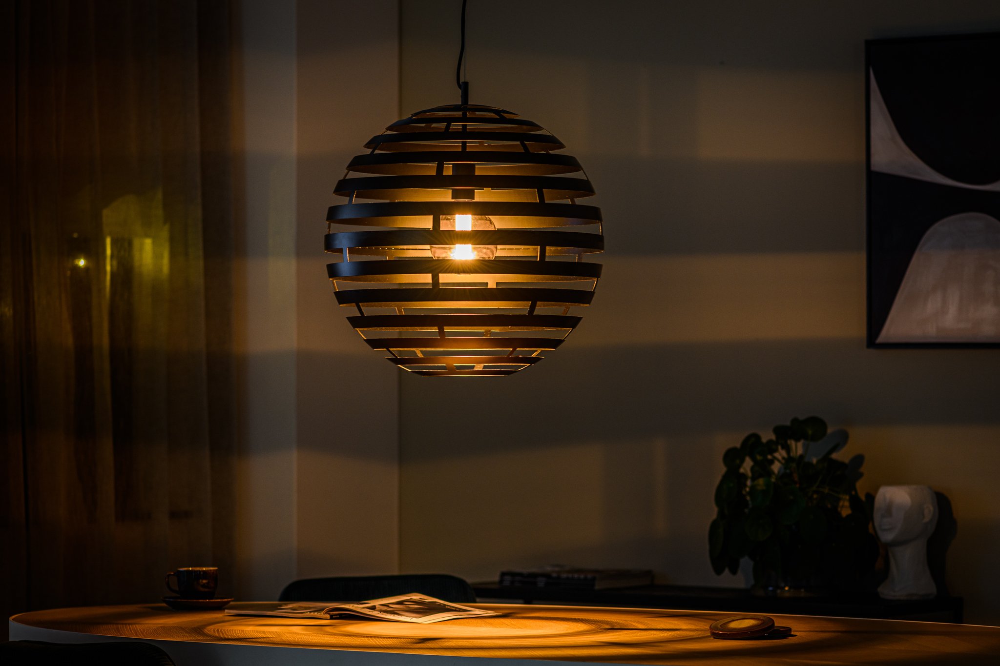 Een bolvormige hanglamp met horizontale lamellen werpt warm, gestructureerd licht op een ronde houten tafel eronder. Een open boek en een kopje staan op tafel, met abstracte kunst aan de muur in een schemerige kamer.