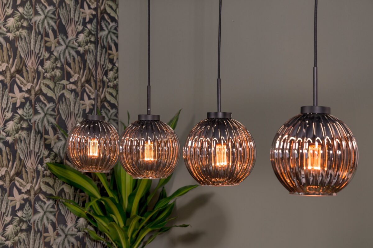 Vier hanglampen met geribbelde glazen kappen hangen op een rij en stralen een warme gloed uit. Ze staan tegen een groene muur met een bladerbehangpatroon en een hoge kamerplant op de achtergrond.