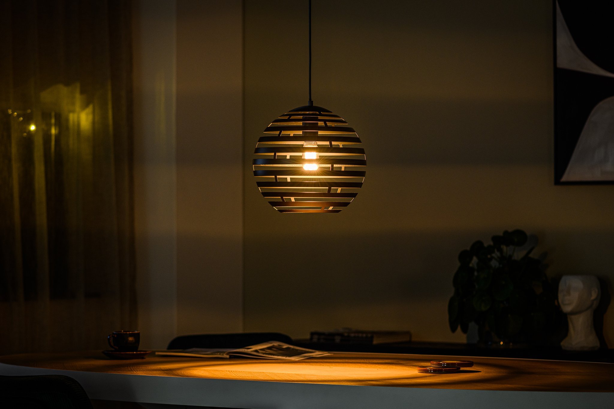 Een warm verlichte moderne eetkamer heeft een bolvormige hanglamp met horizontale lamellen die boven een houten tafel hangt. Op de tafel staan een koffiekopje, een schoteltje en een opengeslagen tijdschrift. Een plant en decoratieve items staan op de achtergrond.