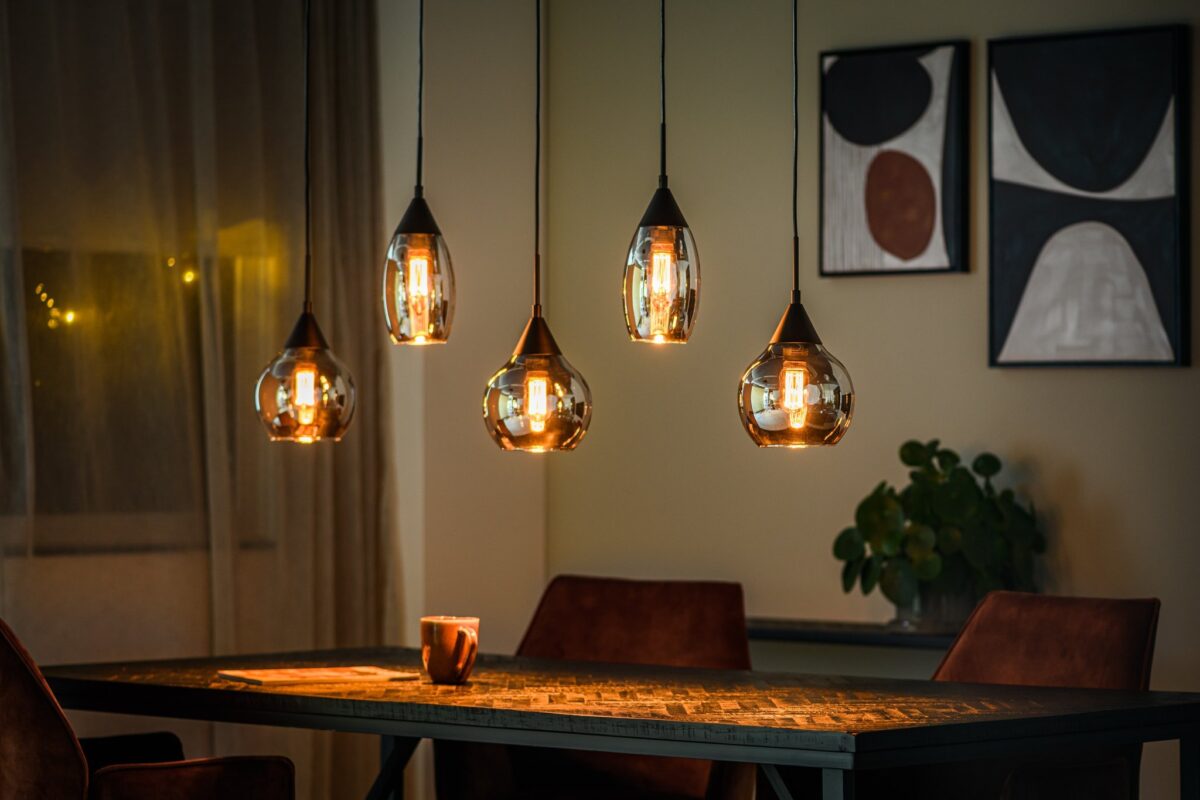 Een gezellige eethoek met vijf hangende glazen hanglampen boven een rechthoekige tafel met een gestructureerd oppervlak. Een mok staat op tafel en abstracte kunst hangt aan de muur. Zachte verlichting en een grote potplant voegen warmte toe aan de scène.