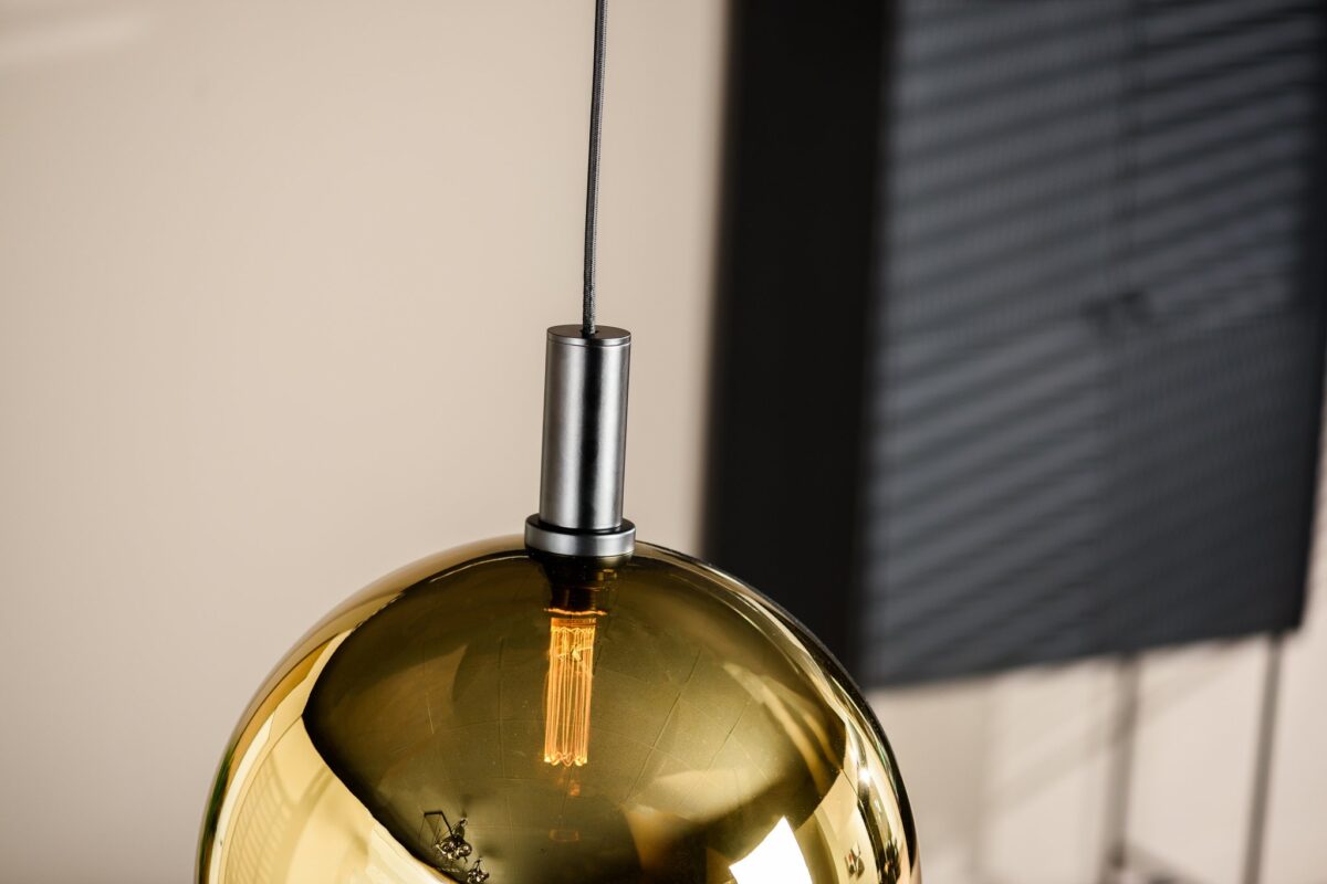 Close-up van een metalen gouden hanglamp met een zichtbare gloeilamp die aan een donkere kabel aan het plafond hangt. De achtergrond heeft vage verticale lijnen, wat doet denken aan een raam of jaloezieën.
