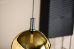 Close-up van een metalen gouden hanglamp met een zichtbare gloeilamp die aan een donkere kabel aan het plafond hangt. De achtergrond heeft vage verticale lijnen, wat doet denken aan een raam of jaloezieën.