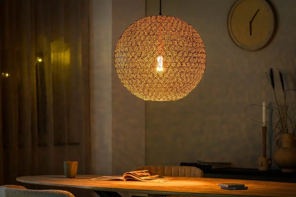 Een gezellige kamer met een gloeiende, getextureerde bolvormige hanglamp boven een houten tafel. Een opengeslagen tijdschrift en een mok rusten op de tafel. Op de achtergrond is er een wandklok en zachte, sfeervolle verlichting.