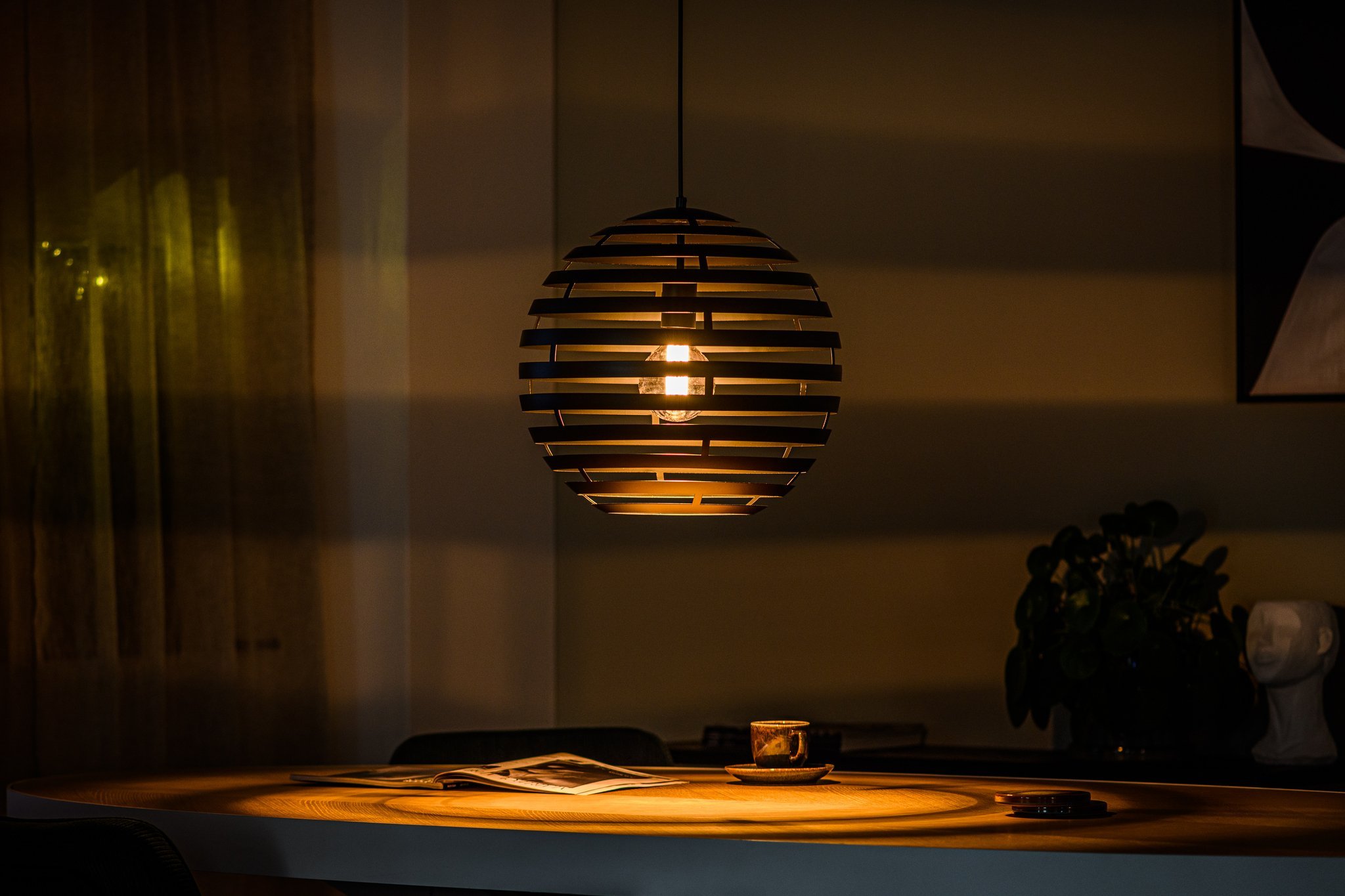 Een schemerige kamer met een moderne bolvormige hanglamp boven een ronde tafel. Op de tafel staan een open boek, een kopje en een klein bordje. Op de achtergrond is een plant te zien die zachte schaduwen werpt.