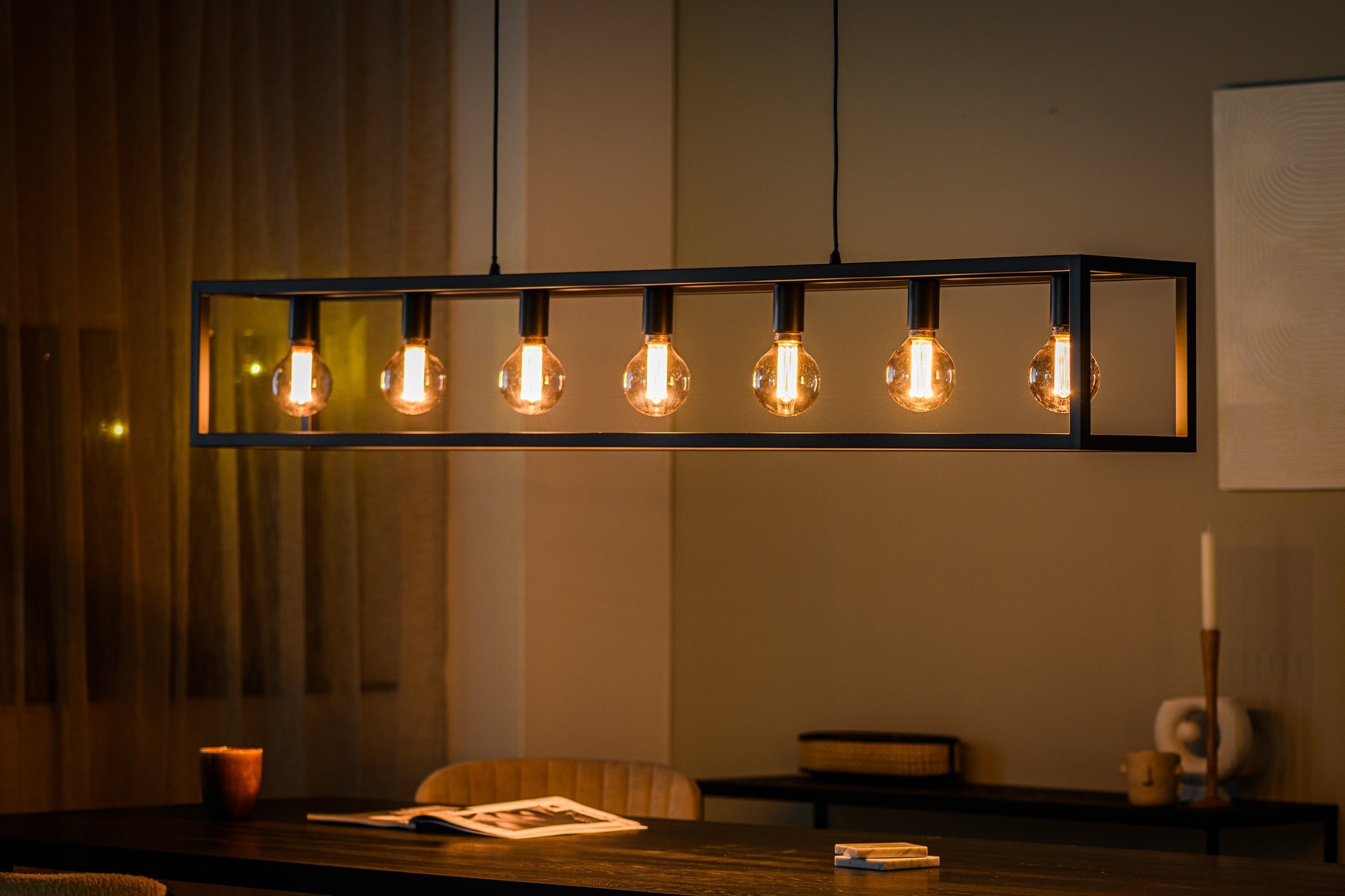 Een moderne rechthoekige kroonluchter met zes ronde, gloeiende lampen hangt boven een eettafel. De kamer heeft een warme ambiance met een kopje op tafel, een tijdschrift en zachte gordijnen op de achtergrond.