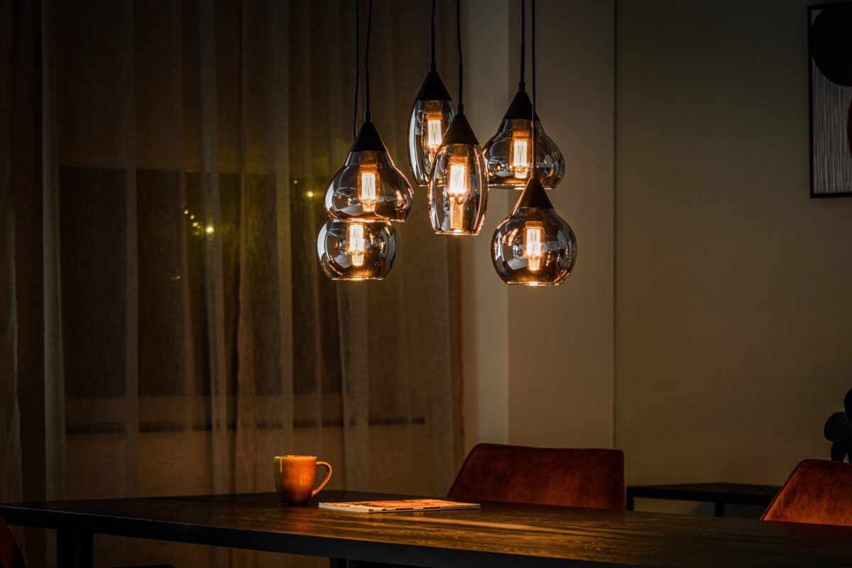 Een schemerige kamer met een cluster van hangende hanglampen boven een donkere tafel. De warme gloed van de lampen creëert een gezellige sfeer. Een mok en een tijdschrift staan op tafel, met een zacht gordijn op de achtergrond.
