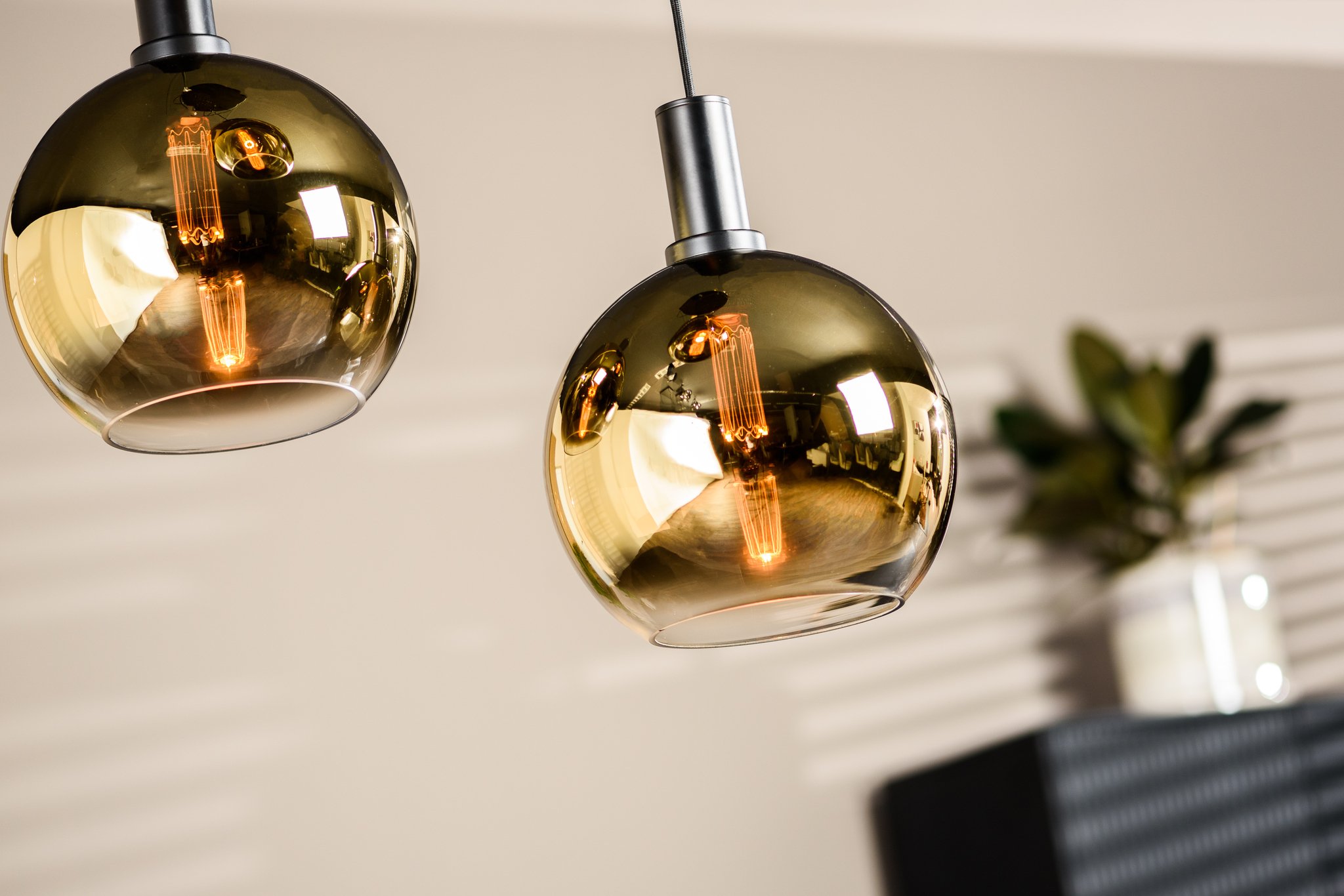Close-up van twee moderne bolvormige hanglampen met een reflecterende gouden afwerking, hangend aan een plafond. Ze zijn verlicht en stralen een warme gloed uit. Op de wazige achtergrond is een kleine potplant zichtbaar.