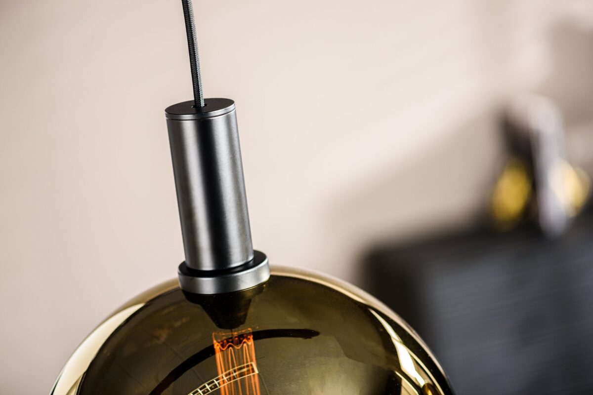 Een close-up van een moderne hanglamp met een zwarte armatuur en een bronskleurige glazen kap. De gloeilamp binnenin is zichtbaar en straalt een warme gloed uit. De achtergrond is zacht gefocust, wat een elegante en eigentijdse ambiance geeft.