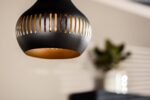Een close-up van een moderne zwarte hanglamp met sleufopeningen, die een warme gloed uitstraalt. De achtergrond is wazig en toont een abstract decor met planten en een wit en zwart kleurenschema.