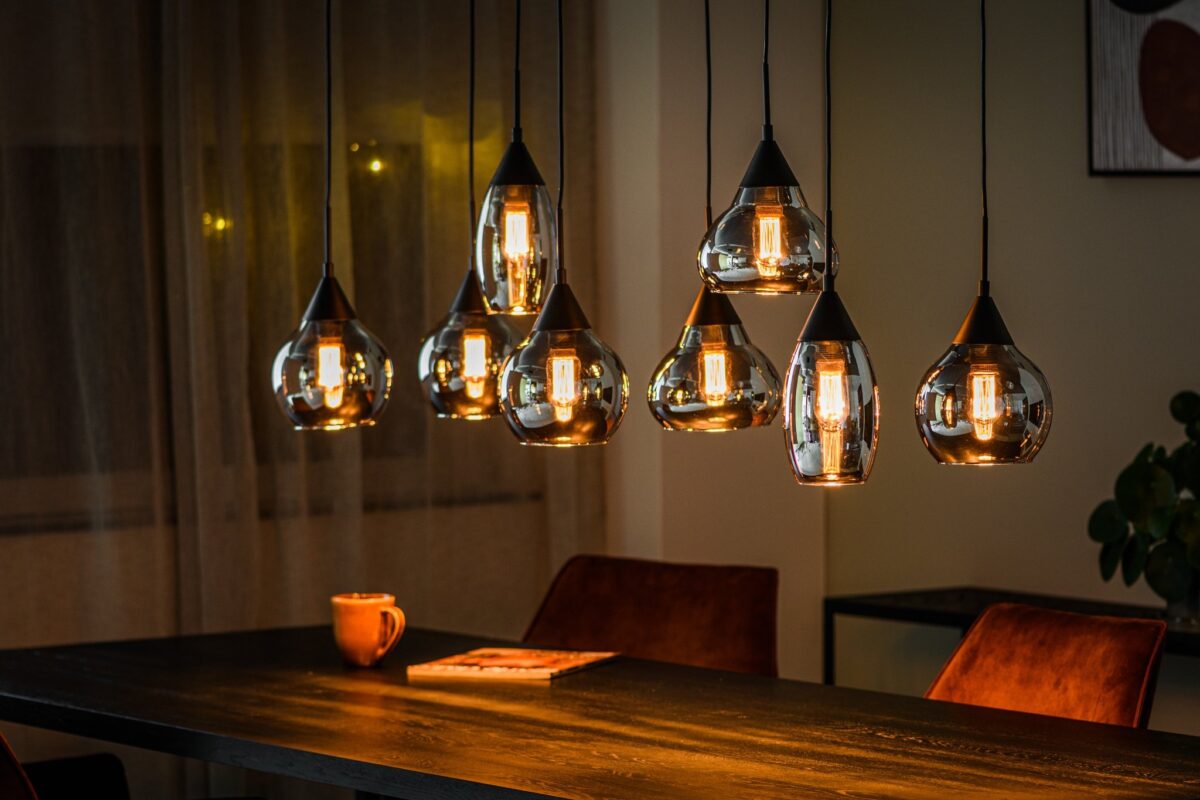 Een eethoek met meerdere moderne hanglampen boven een donkere houten tafel. De warme gloed verlicht een kopje en een boek op de tafel, met twee bruine gestoffeerde stoelen gedeeltelijk zichtbaar. Gordijnen en een plant zijn op de achtergrond.