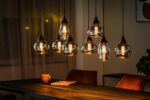 Een eethoek met meerdere moderne hanglampen boven een donkere houten tafel. De warme gloed verlicht een kopje en een boek op de tafel, met twee bruine gestoffeerde stoelen gedeeltelijk zichtbaar. Gordijnen en een plant zijn op de achtergrond.