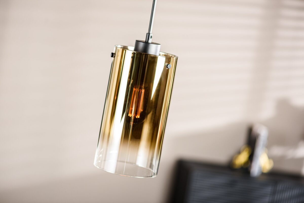Een moderne cilindrische hanglamp met een gouden metalen afwerking, hangt aan het plafond. Het licht is aan en werpt een warme gloed. Vage achtergrond met horizontale schaduwen op een beige muur en een donker meubelstuk.