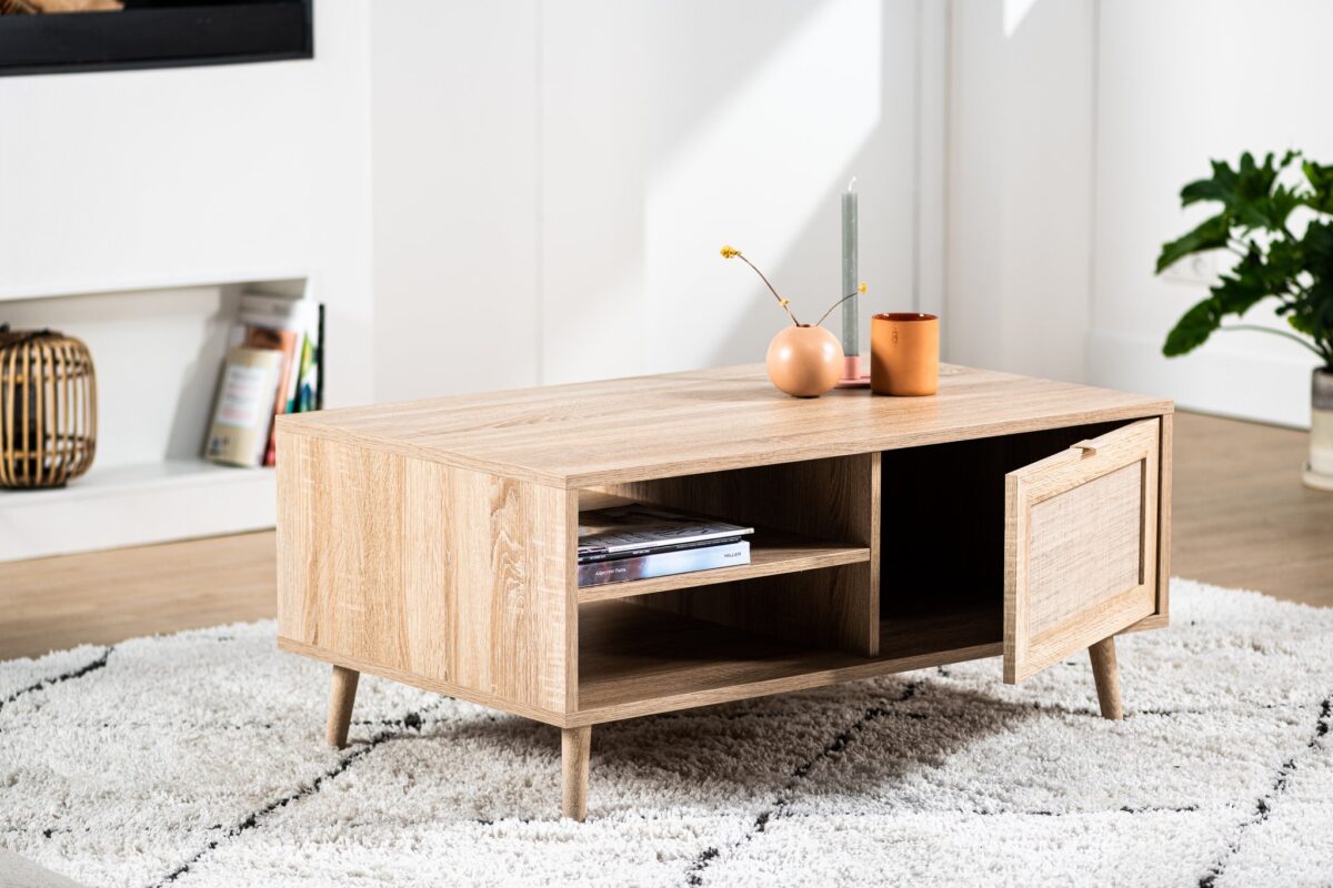 De Salontafel, 100x60 cm, M930 Sedona met schuine poten staat op een tapijt met witte structuur. Het heeft open planken met boeken, een deur op een kier en is bedekt met decoratieve items en een kaars. Een plant decoreert de achtergrond.