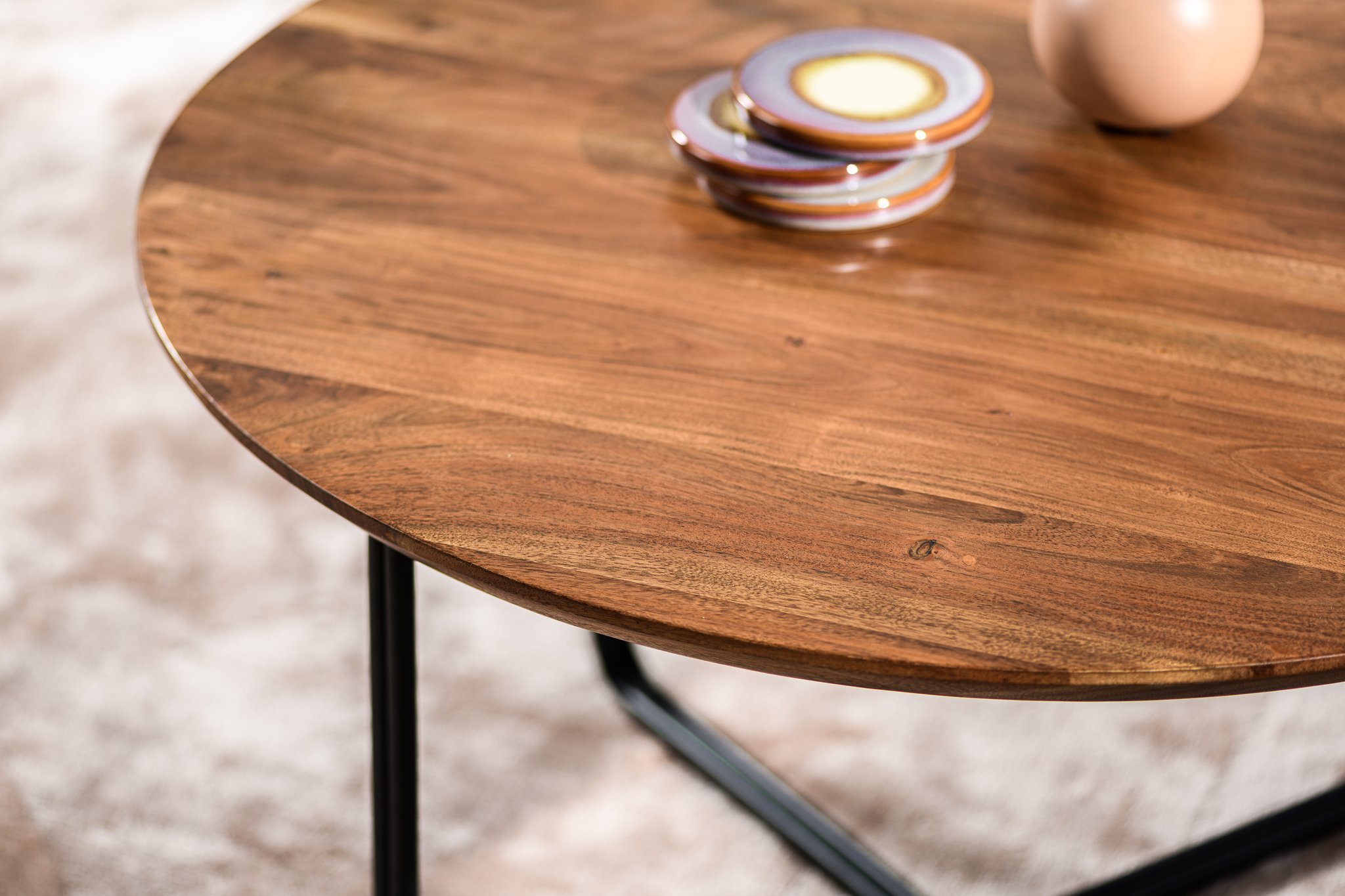 Een close-up van de Salontafel rond, 75 cm, B430 bruin toont het gladde ronde houten blad met gestapelde keramische borden en een vaas, ondersteund door zwart metalen poten tegen een zacht vervaagde achtergrond.