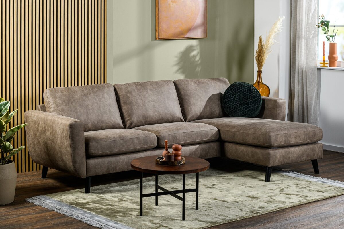 Een moderne woonkamer is voorzien van de 3 zits bank CL L+R in Savannah S520 taupe stof, een kleine ronde houten salontafel, een lichtgroen vloerkleed, potplanten, vensterbankdecoratie, verticale houten lambrisering en ingelijste muurkunst.