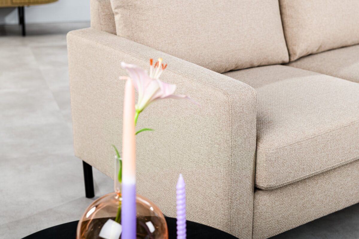 Een close-up van de 3 zits bank CL L+R in Dillon D460 beige stof met een zwarte ronde salontafel met daarop een glazen vaas met een roze lelie en twee ombre kaarsen, op een lichtgrijze vloer.