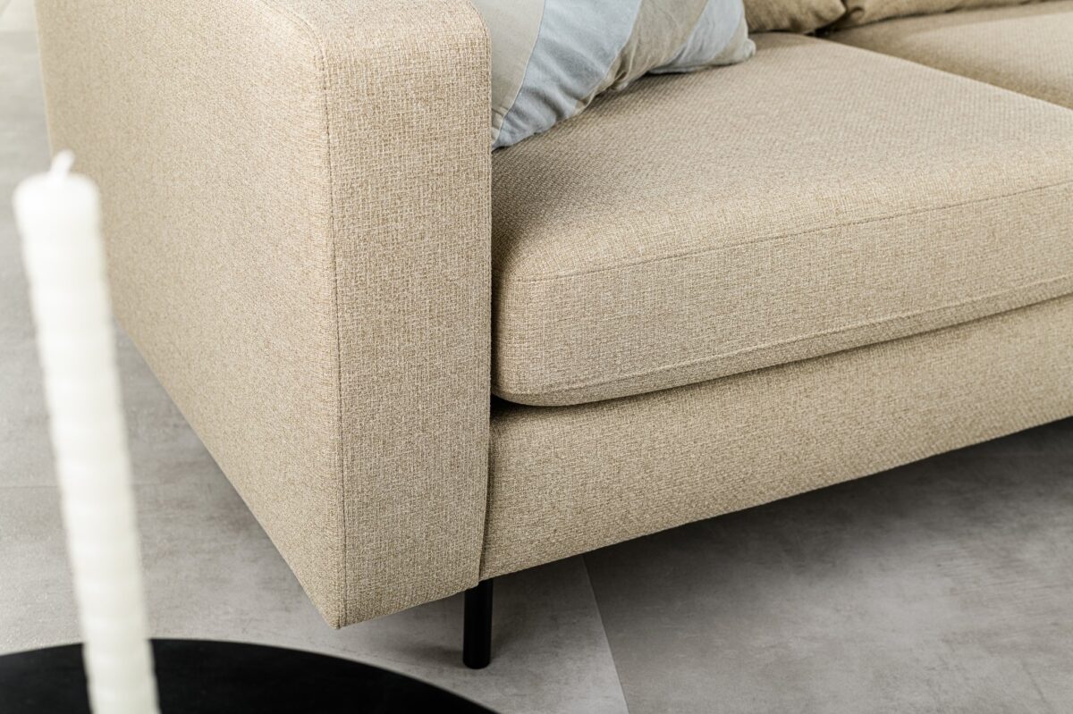 Close-up van de 3 zits bank CL L+R in D460 beige Dillon stof met zwarte poten op een lichte vloer. Een lichtblauw en grijs gestreept kussen rust op de bank, terwijl een deel van een zwarte ronde tafel en een witte kaars op de voorgrond verschijnen.