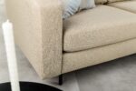 Close-up van de 3 zits bank CL L+R in D460 beige Dillon stof met zwarte poten op een lichte vloer. Een lichtblauw en grijs gestreept kussen rust op de bank, terwijl een deel van een zwarte ronde tafel en een witte kaars op de voorgrond verschijnen.