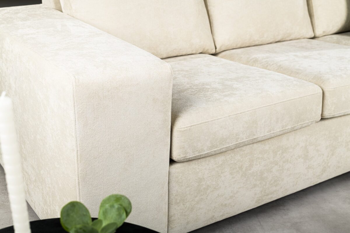Close-up van een lichtbeige, getextureerde bank met een brede armleuning en zachte kussens. Een kleine zwarte tafel met een groene plant is gedeeltelijk zichtbaar op de voorgrond. De setting oogt modern en minimalistisch.