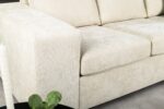 Close-up van een lichtbeige, getextureerde bank met een brede armleuning en zachte kussens. Een kleine zwarte tafel met een groene plant is gedeeltelijk zichtbaar op de voorgrond. De setting oogt modern en minimalistisch.
