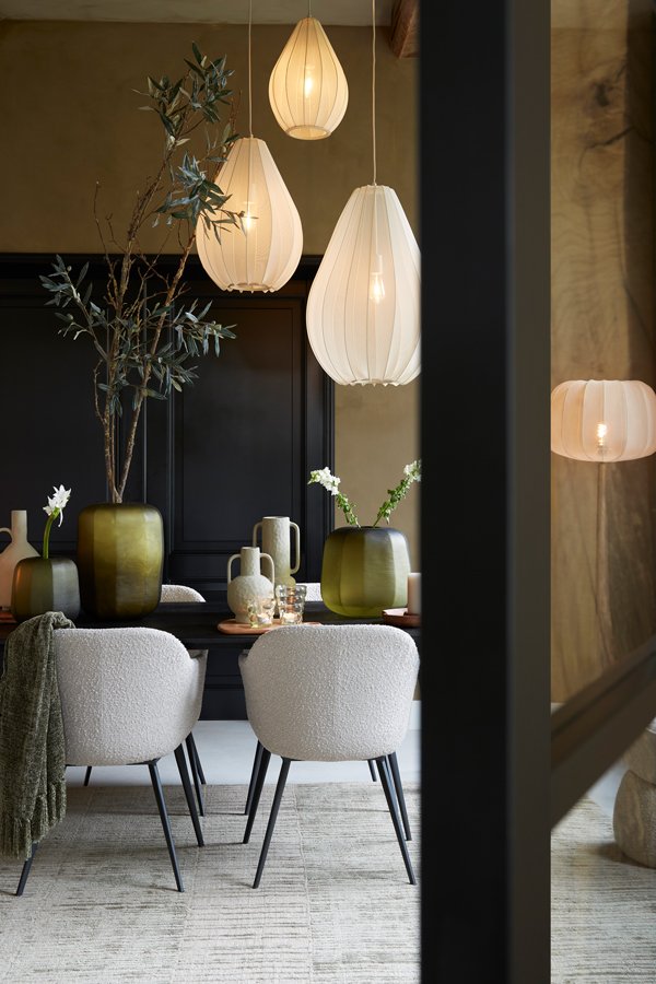 Moderne eetkamer met een houten tafel, witte stoelen met kussens, groene vazen en decoratieve takken. Drie grote, witte, hangende hanglampen verlichten de ruimte. De kamer heeft een warm en minimalistisch design, met neutrale tinten en zachte texturen.