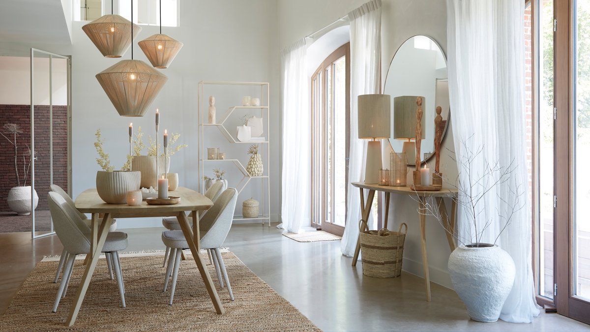 Een moderne eetkamer met een houten tafel en grijze stoelen. Hanglampen hangen erboven en kaarsen staan op tafel. Een grote spiegel en lamp staan op een consoletafel bij de boogramen met doorschijnende gordijnen. Neutrale decoratie accentueert de ruimte.