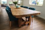 Een rustieke houten eettafel met een dikke, natuurlijke afwerking wordt omringd door zes blauwgroene gestoffeerde stoelen. De tafel is versierd met kaarsen en een bloemstuk. Zonlicht filtert door een raam en verlicht de houten vloer.
