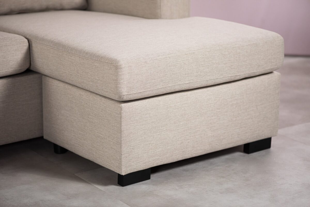 Close-up van een beige gestoffeerde ottoman-stijl sectie van een bank met een lichte stoffen textuur. Het heeft een rechthoekige vorm met zwarte poten, geplaatst op een gladde, lichtgekleurde vloer.