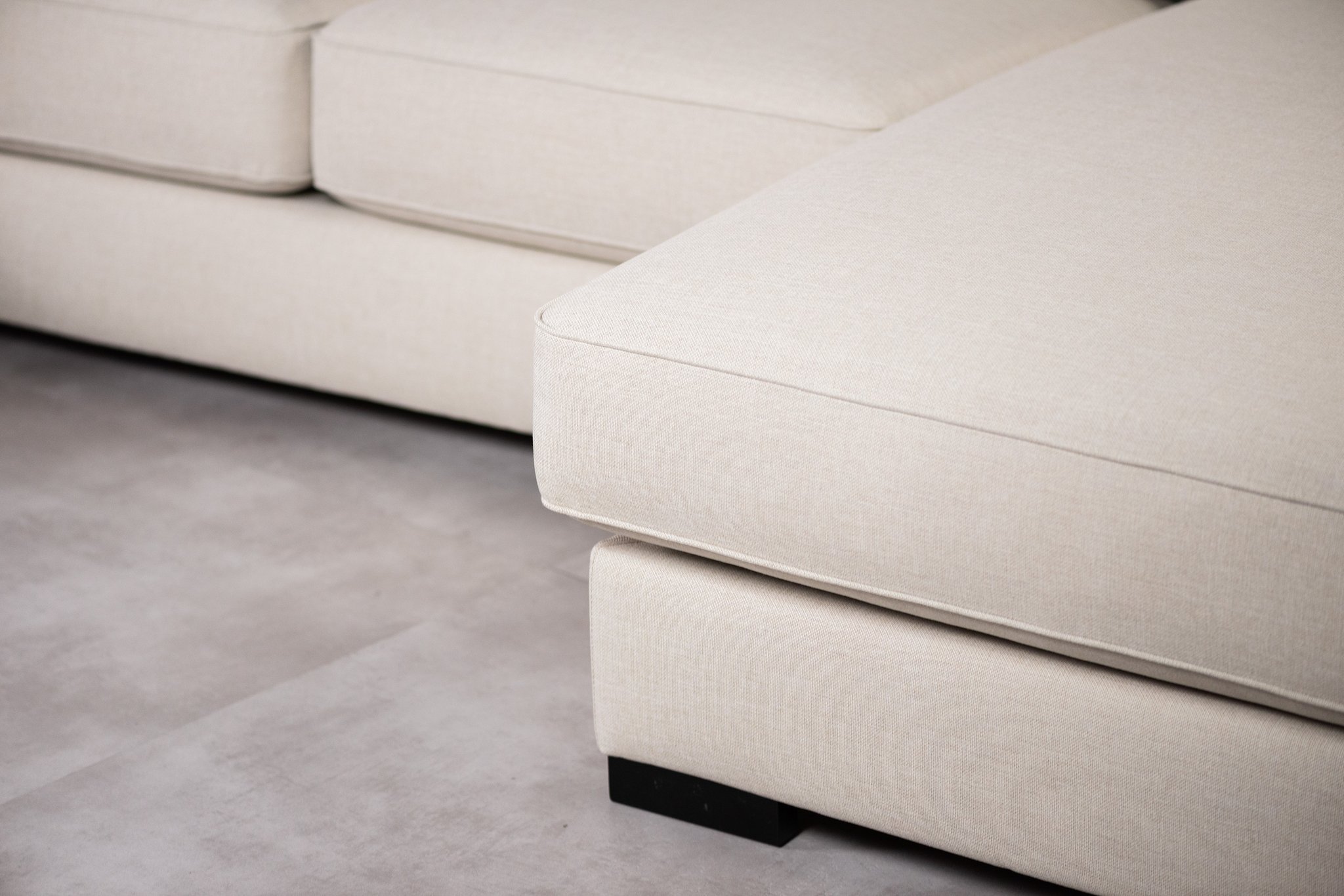 Close-up van een beige sectionele bank met zichtbare stiksels op een lichtgrijze vloer. De afbeelding benadrukt de strakke lijnen en textuur van de stoffen bekleding.