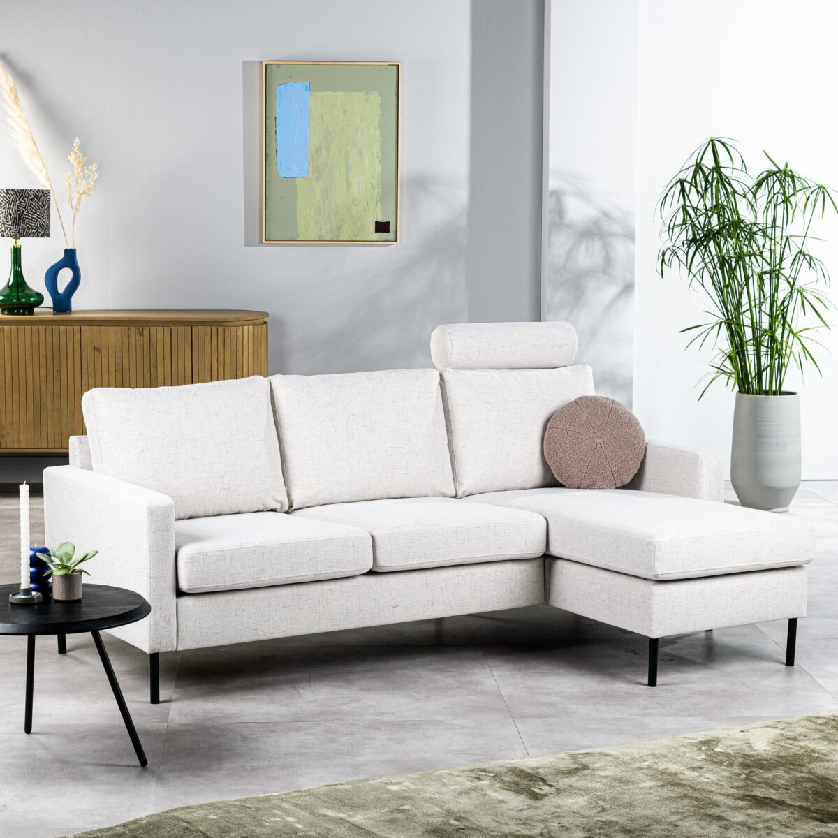 Een moderne woonkamer met een witte hoekbank met een rond kussen. Er staat een zwarte bijzettafel met kaarsen en een kleine plant ernaast. Een houten dressoir met decoratie-items staat tegen de muur en er staat een hoge potplant in de buurt.