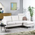 Een moderne woonkamer met een witte hoekbank met een rond kussen. Er staat een zwarte bijzettafel met kaarsen en een kleine plant ernaast. Een houten dressoir met decoratie-items staat tegen de muur en er staat een hoge potplant in de buurt.