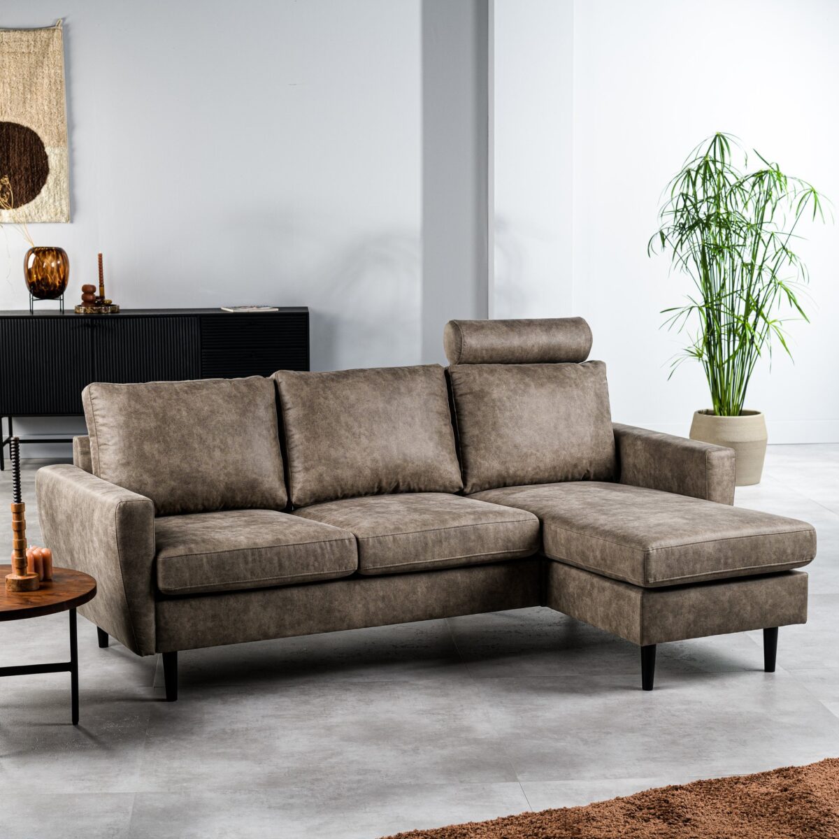 Een moderne woonkamer met een bruine L-vormige bank met zwarte poten. Een hoge groene plant staat naast de bank. De kamer heeft een zwart dressoir met decoratieve items en een wandkleed. De vloer is lichtgrijs en een bruin kleed is gedeeltelijk zichtbaar.