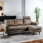 Een moderne woonkamer met een bruine L-vormige bank met zwarte poten. Een hoge groene plant staat naast de bank. De kamer heeft een zwart dressoir met decoratieve items en een wandkleed. De vloer is lichtgrijs en een bruin kleed is gedeeltelijk zichtbaar.