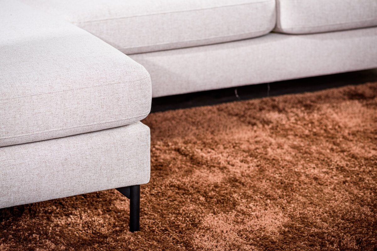 Close-up van een lichtbeige sectionele bank op een pluche, getextureerd bruin kleed. De bank heeft zwarte poten en ziet er modern en comfortabel uit, met strakke lijnen. Het getoonde gedeelte van de bank bevat een hoek met een verlengde zitting.