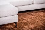 Close-up van een lichtbeige sectionele bank op een pluche, getextureerd bruin kleed. De bank heeft zwarte poten en ziet er modern en comfortabel uit, met strakke lijnen. Het getoonde gedeelte van de bank bevat een hoek met een verlengde zitting.