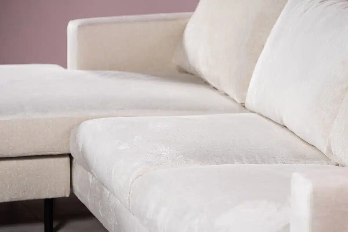 Close-up van een beige, getextureerde sectionele bank met een chaise longue op een lichtgekleurde vloer. De bank heeft strakke, geometrische lijnen en staat tegen een subtiele roze muur, wat een zachte, uitnodigende sfeer creëert.