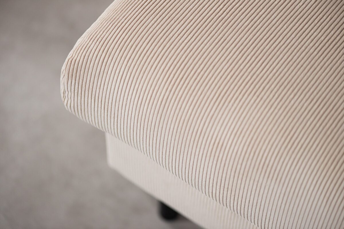 Close-up van een beige stof met een getextureerd, geribbeld patroon dat een meubelstuk bedekt, mogelijk een armleuning van een bank of een stoel. De zachte, neutrale kleur en fijne lijnen creëren een subtiele, elegante uitstraling.