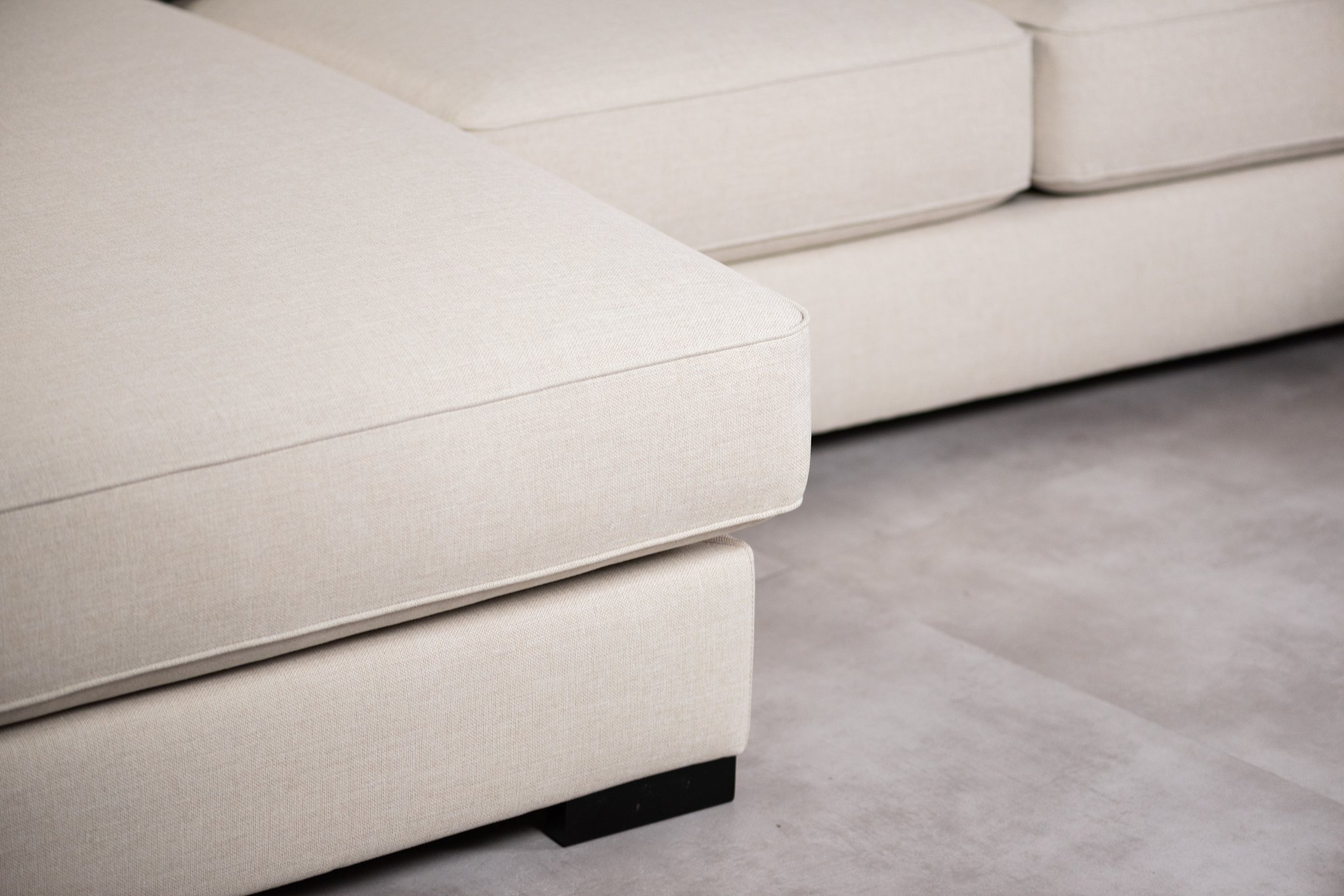 Close-up van een lichtbeige sectionele bank op een grijze vloer. De afbeelding toont twee zachte kussens met een minimalistisch en modern design. De textuur van de stof is glad en de strakke lijnen van het meubilair creëren een eigentijdse look.