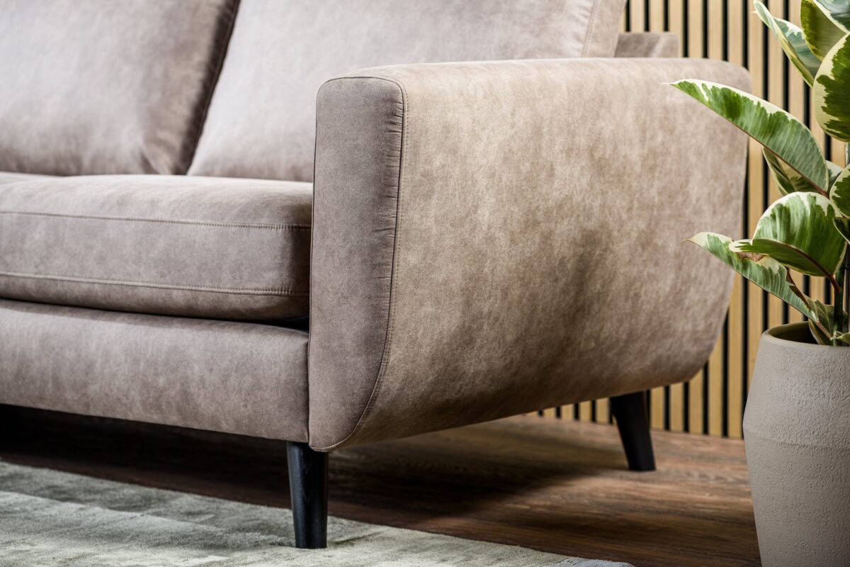 Close-up van de 3 zits bank CL L+R in Savannah S520 taupe stof met zwarte poten op een houten vloer, naast een groene potplant. Lichte houten latten vormen de achtergrondmuur en een licht vloerkleed is gedeeltelijk zichtbaar.