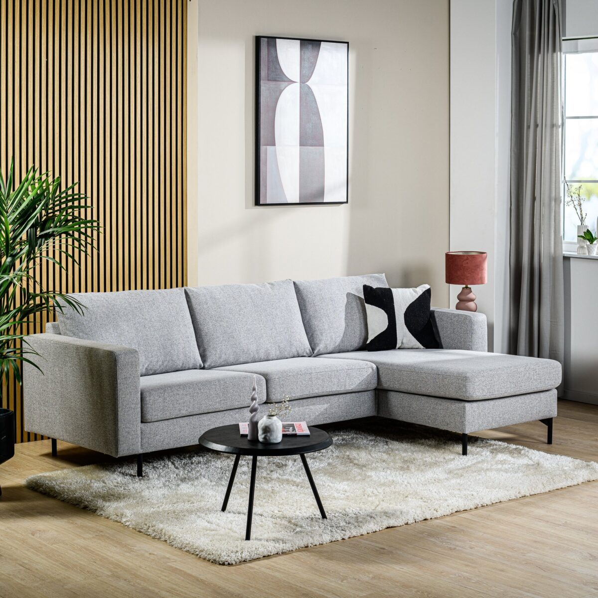 Een moderne woonkamer met de 3 zits bank CL L+R in de stof Chloe C311 grijs op een wit vloerkleed, gecombineerd met een ronde zwarte salontafel, abstracte muurkunst, een rode tafellamp, een raam met gordijnen en een groene plant in de hoek.