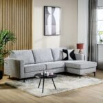 Een moderne woonkamer met de 3 zits bank CL L+R in de stof Chloe C311 grijs op een wit vloerkleed, gecombineerd met een ronde zwarte salontafel, abstracte muurkunst, een rode tafellamp, een raam met gordijnen en een groene plant in de hoek.