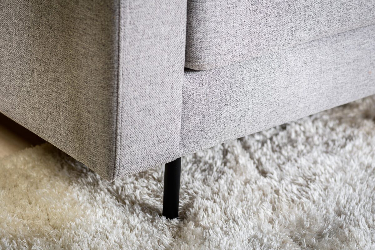 Close-up van de hoek van de 3-zits bank CL L+R in stof Chloe C311 grijs, met zwarte metalen poten en geplaatst op een dik off-white shaggy tapijt.