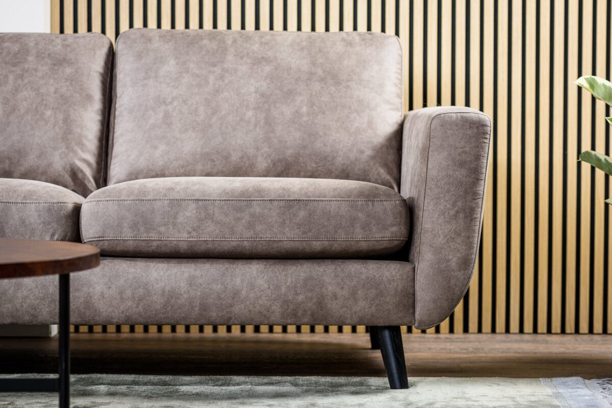 Een close-up van de 3 zits bank CL L+R in de stof Savannah S520 taupe, met pluche kussens en zwarte poten, tegen een wand met houten latten. Een deel van een salontafel en een bladplant zijn ook gedeeltelijk zichtbaar.