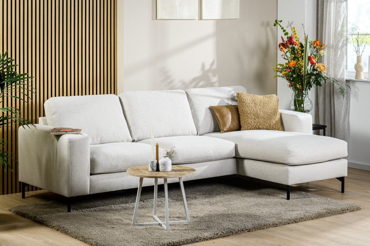 Een moderne woonkamer met een lichtbeige hoekbank op een beige kleed. Er staat een ronde houten salontafel met kaarsen, een bruin kussen en sprei op de bank, een vaas met kleurrijke bloemen en een houten accentmuur met planten eromheen.