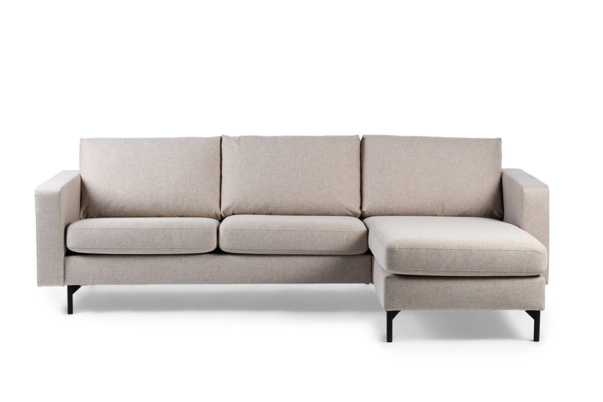 Een moderne, lichtbeige sectionele bank met drie kussens en een chaise longue aan de rechterkant. De bank heeft rechte armleuningen en zwarte metalen poten op een witte achtergrond.