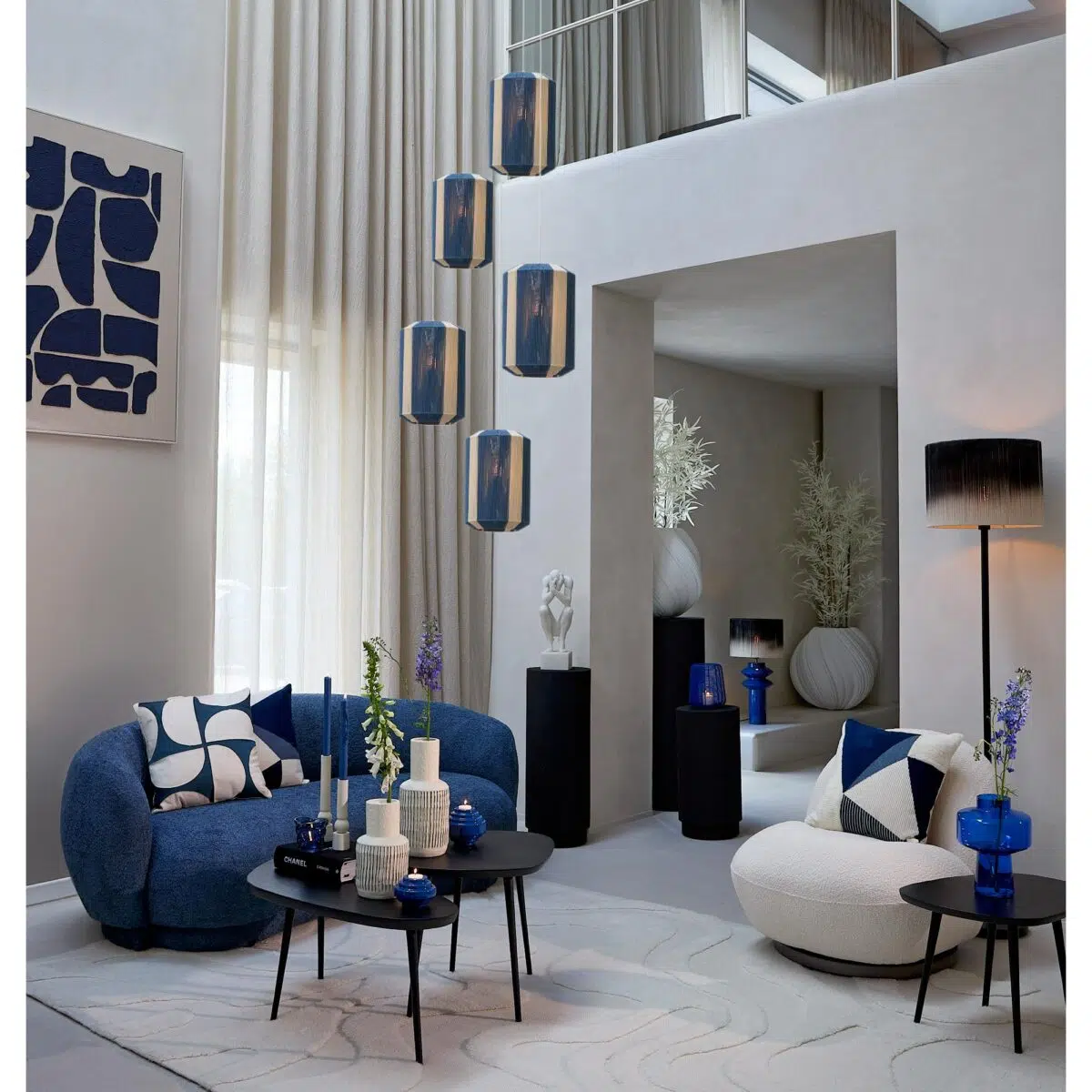 Moderne woonkamer met een blauw en wit thema. Een blauwe bank, witte stoel met kussens en zwarte tafels met geometrische kussens en vazen. Abstracte kunst aan de muur en hangende blauwe hanglampen dragen bij aan het decor. Grote ramen met doorschijnende gordijnen zorgen voor natuurlijk licht.