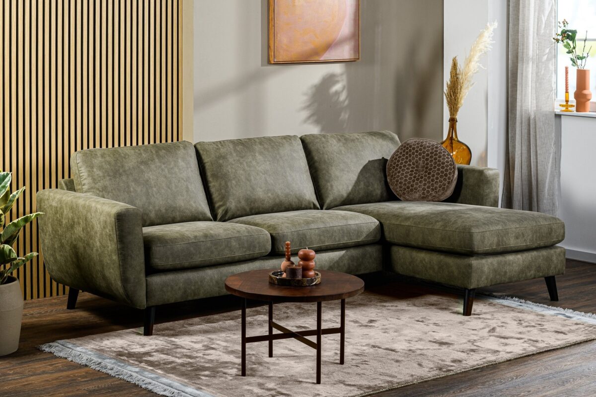Een moderne woonkamer met de 3 zits bank CL L+R in Savannah S156 groen, een ronde houten salontafel, een vloerkleed met structuur, planten, decoratieve vazen en muurkunst in neutrale tinten bij een raam met witte gordijnen.