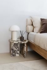 Een minimalistische slaapkamer heeft een houten bed met beige beddengoed en bruine kussens. Een klein marmeren bijzettafeltje met een witte lamp, een zwarte rietdiffuser en een klein schaaltje, allemaal tegen een witte muur en beige vloer.