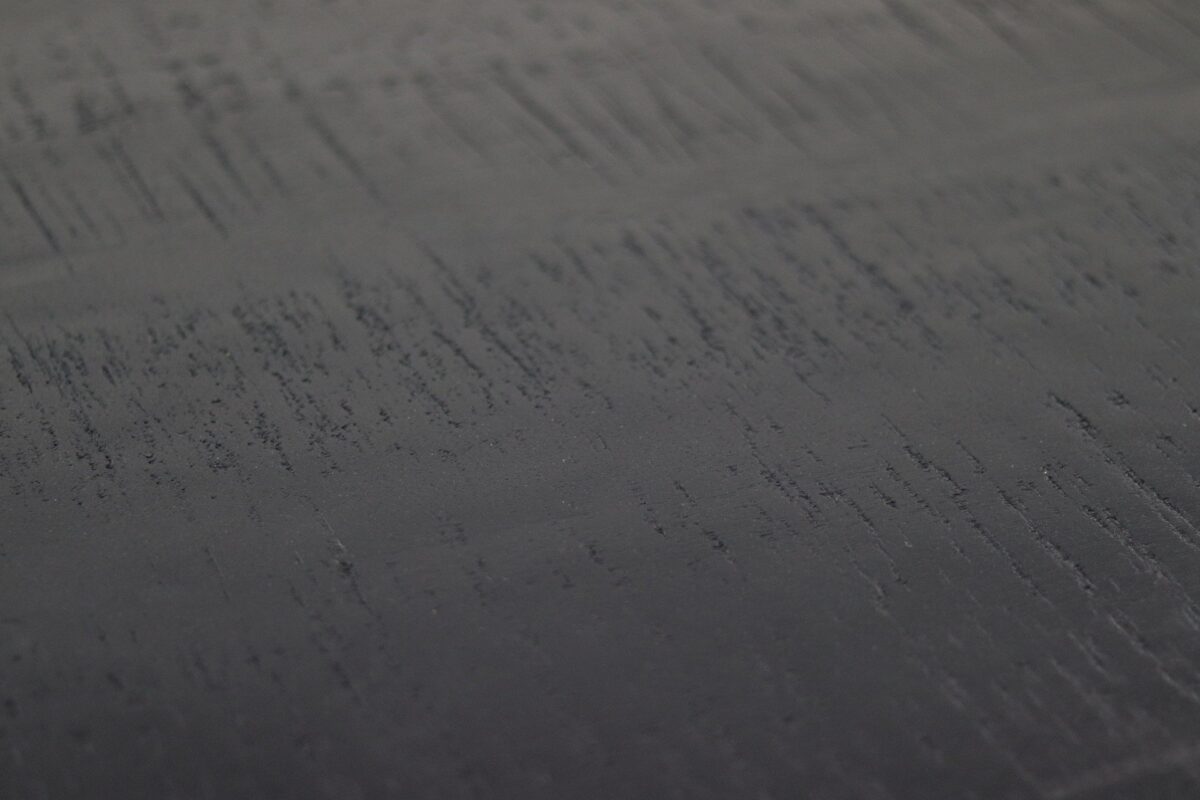 Close-up van een donker, gestructureerd oppervlak met horizontale strepen en subtiele variaties in schaduw, dat lijkt op geborsteld metaal of een licht versleten oppervlak. De textuur creëert een gevoel van diepte en beweging over de afbeelding.