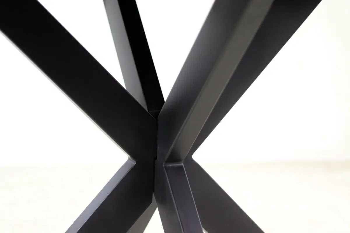 Close-up van een moderne, geometrische zwarte metalen tafelvoet met kruisende, schuine balken die een X-vorm vormen tegen een effen witte achtergrond.